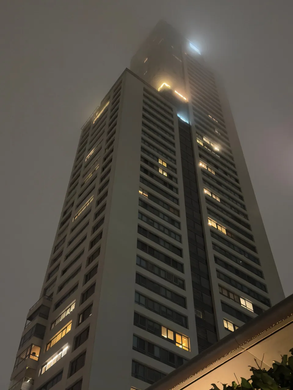 Torre iluminada de noche perdiéndose en la niebla, con ventanas encendidas salteadas